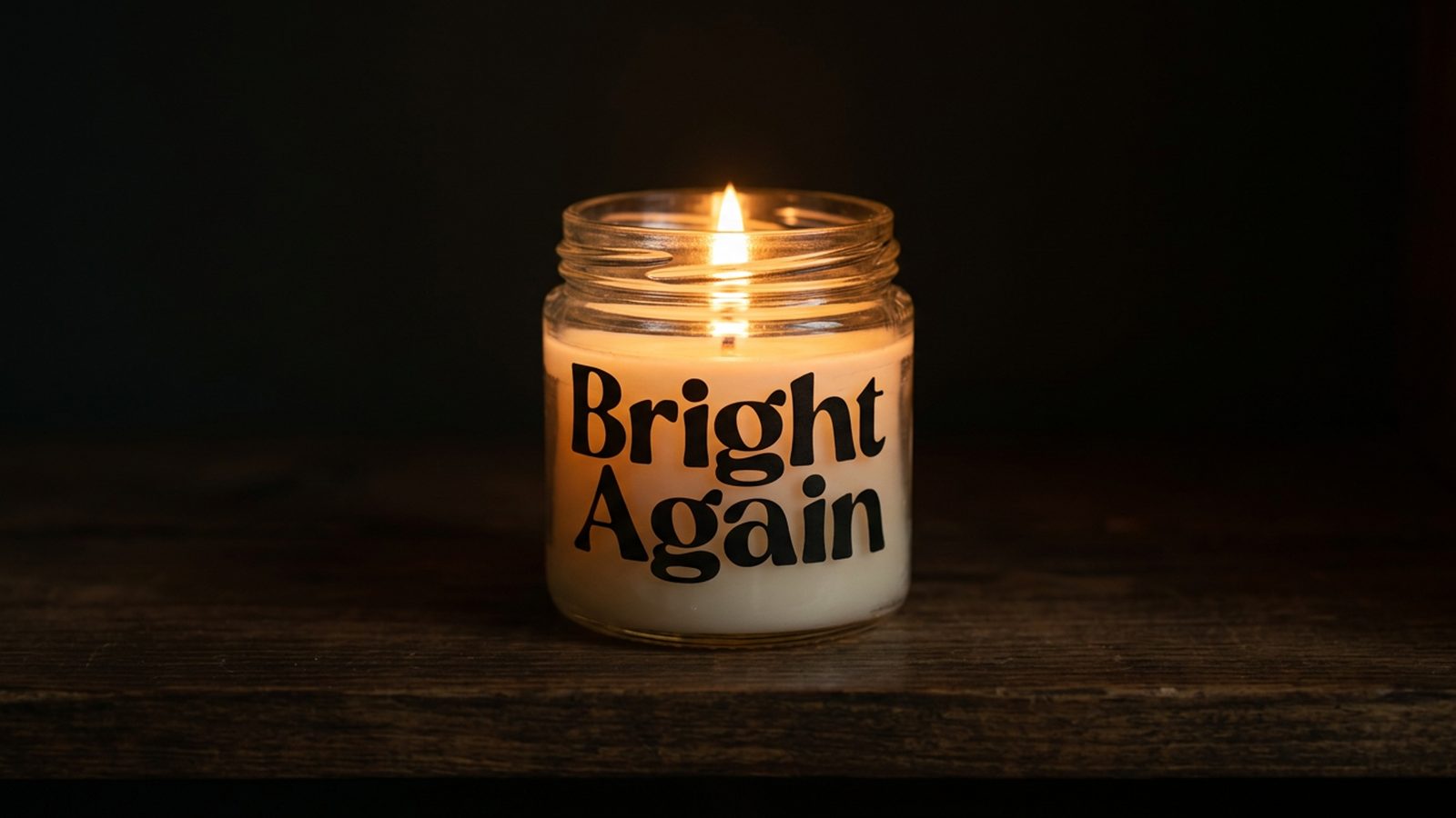 A lit candle in a recycled glass jar glowing warm amber on a dark wood shelf at evening
