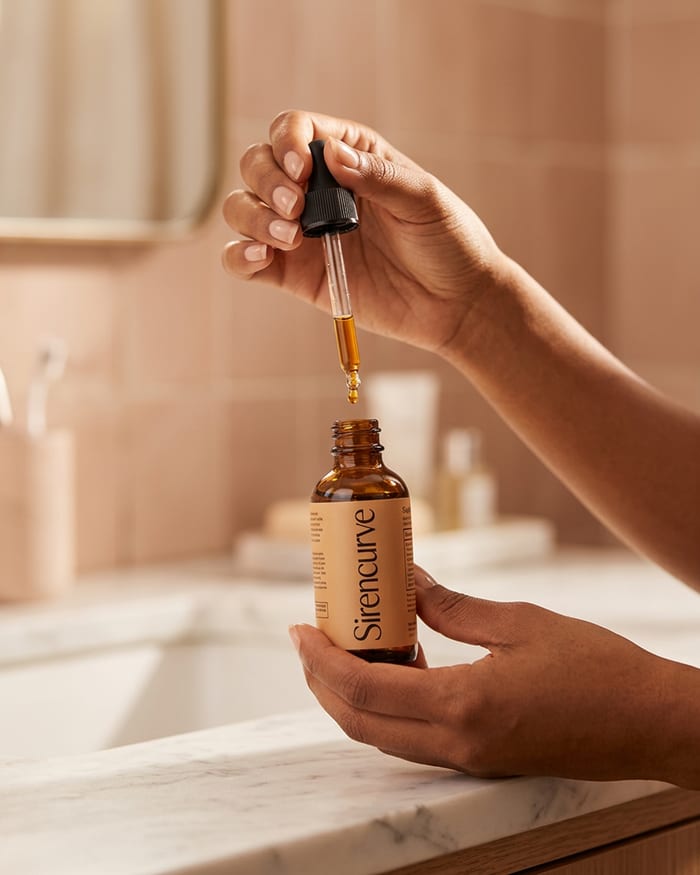 A hand lifting a Sirencurve amber glass dropper bottle, framed in soft morning light — the daily ritual.