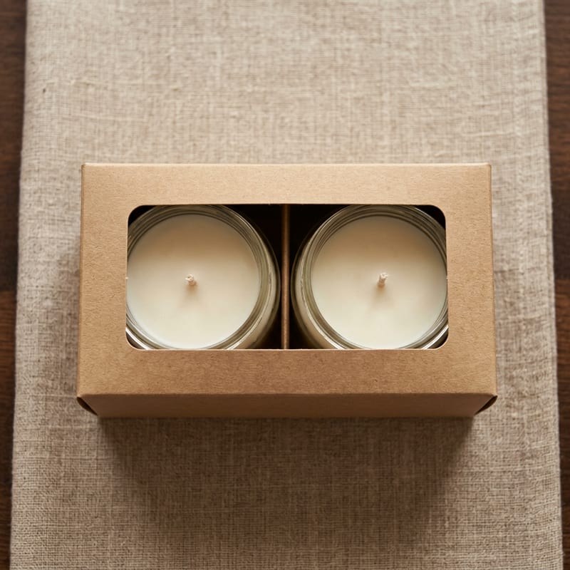 Two Bright Again Signature Candles nestled side by side in a natural kraft paper gift sleeve on raw linen