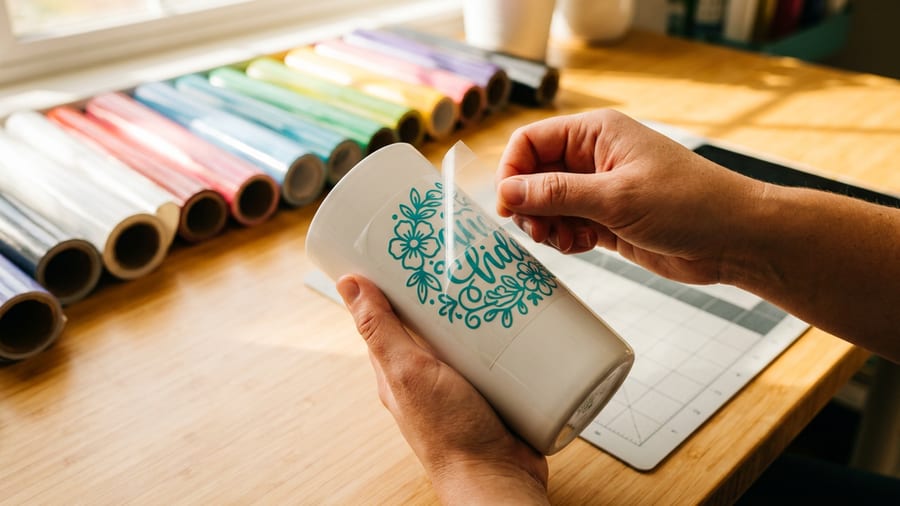 Permanent vinyl rolls fanned out beside finished decals applied to a ceramic tumbler and glass jar on a light wood surface