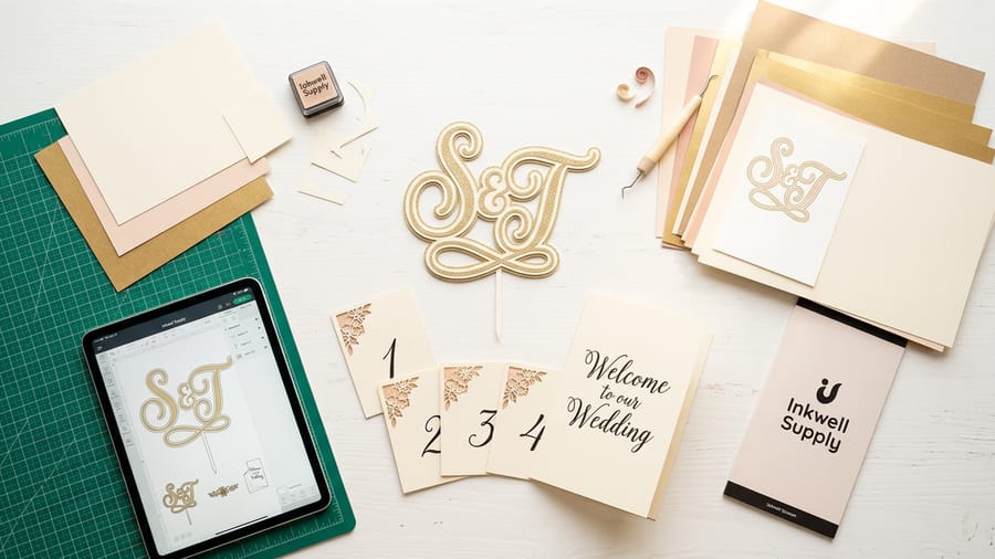 Finished wedding papercraft items including a monogram cake topper, table number cards, and a welcome sign beside a cutting mat and cardstock