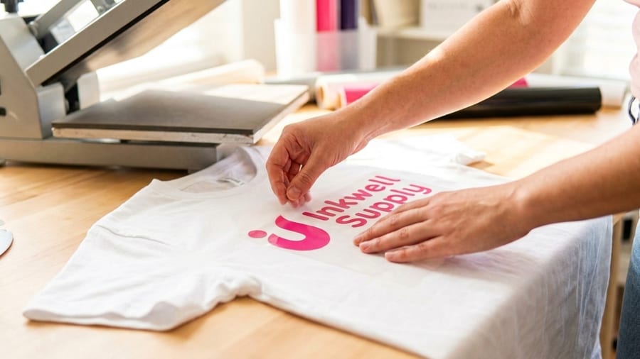 Heat transfer vinyl supplies laid out with finished custom shirts, a cutting machine, and HTV rolls in bold colors