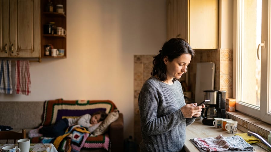 Mamă singură în bucătărie privind telefonul dimineața, copil bolnav odihnindu-se pe canapea în fundal, lumină naturală caldă