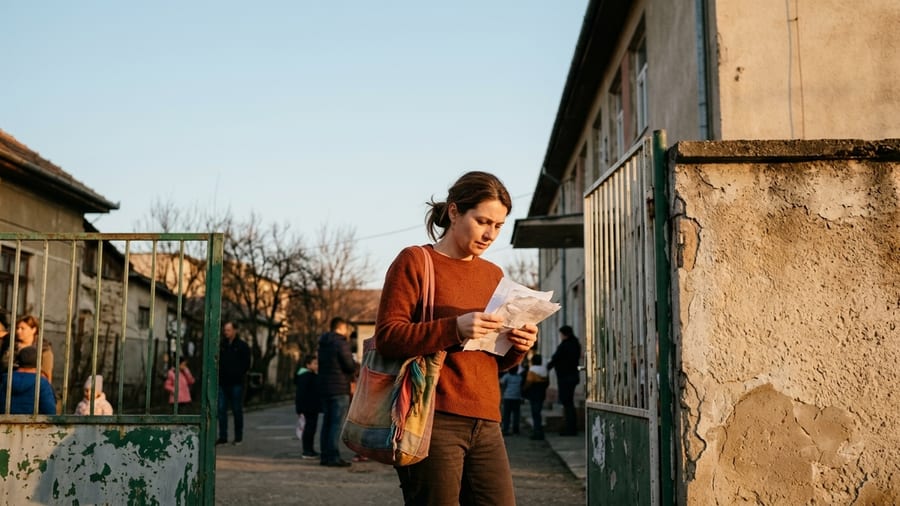 O mamă singură la poarta unei școli românești, ținând câteva acte în mână, lumină caldă de după-amiază
