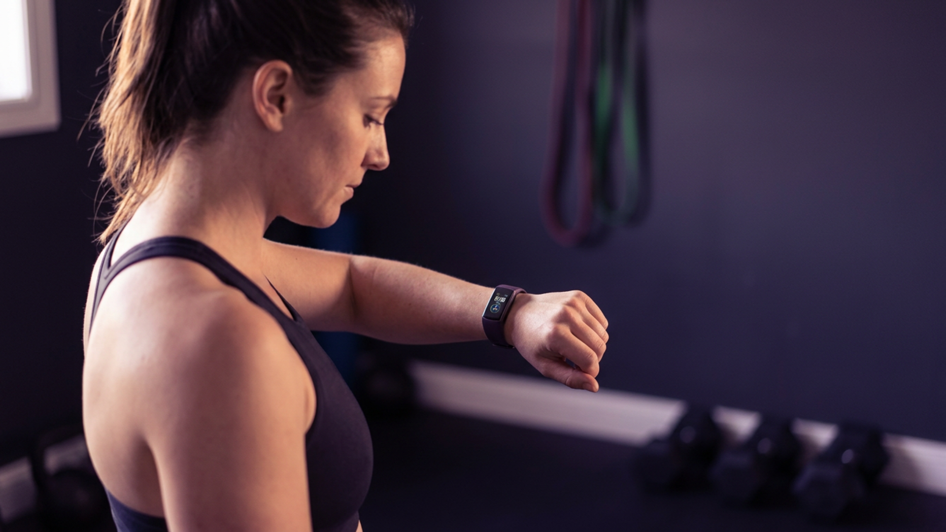 A woman checks her fitness band in a dimly lit home gym at golden hour, the band displaying a health metric on her wrist